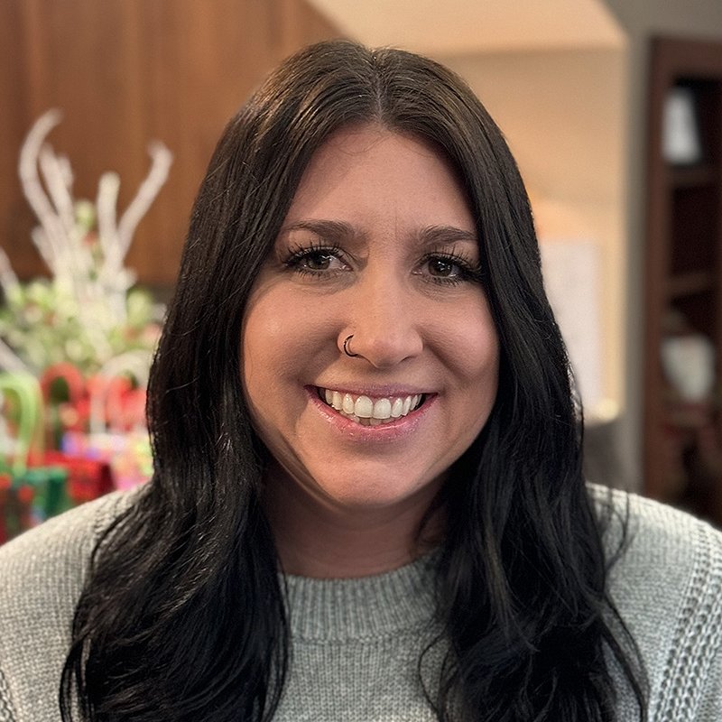 Smiling woman with long dark hair and a nose ring looking at the camera indoors.