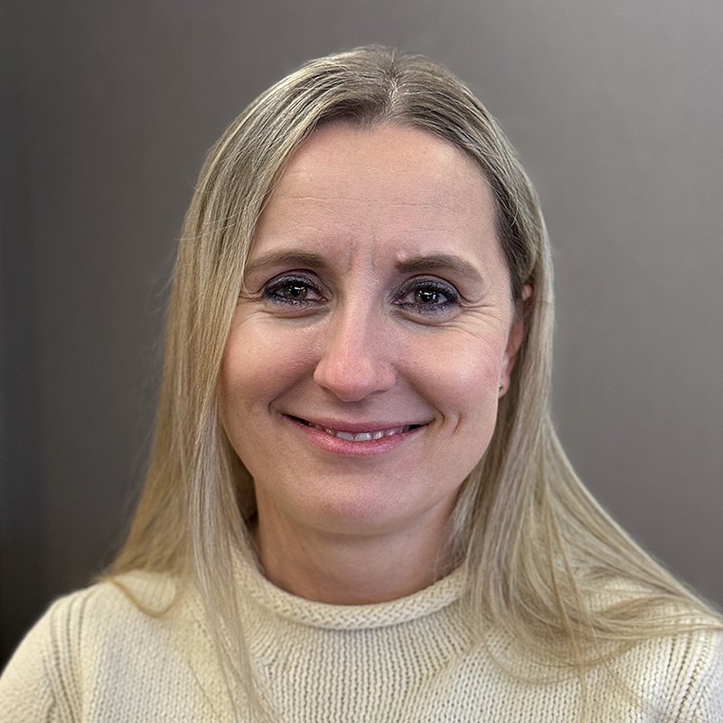 Portrait of a smiling blonde woman in a cream knit sweater against a gray background.