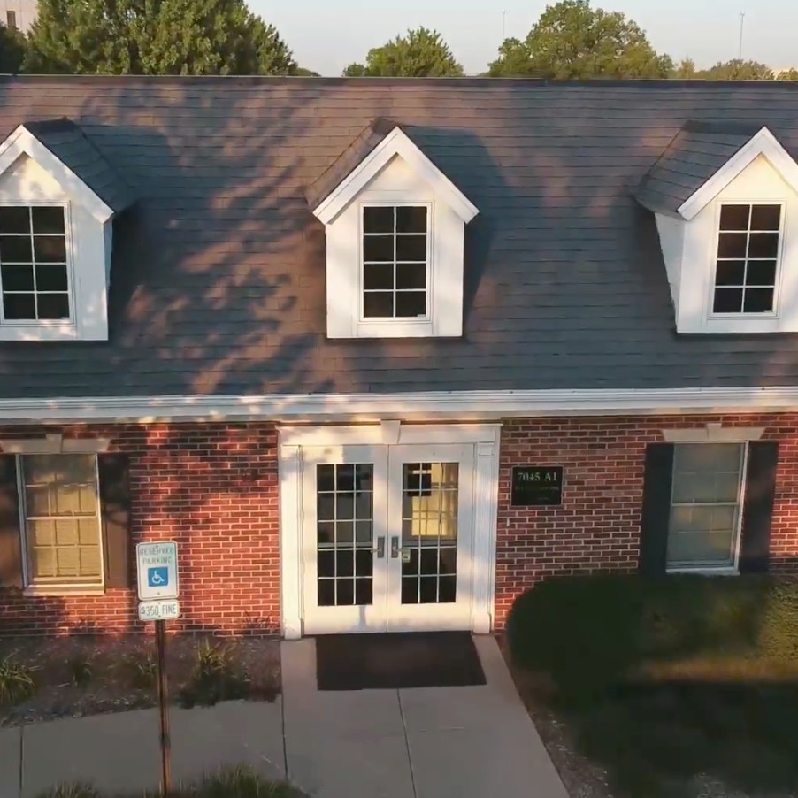 Front entrance of a brick building with double glass doors and dormer windows on the roof; handicap parking sign visible nearby.